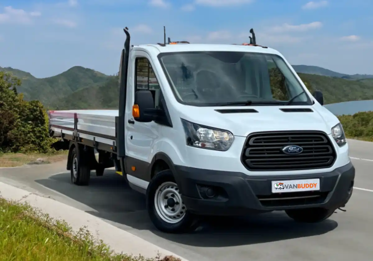 a white Ford Transit Dropside is driving down a road near a lake.